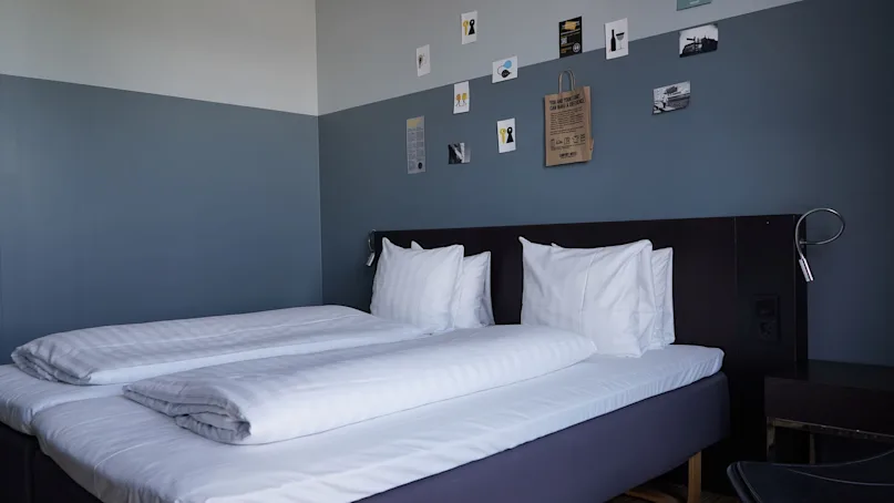 Double bed with white linens against a dual-tone blue wall, adorned with small art pieces. Text on a paper bag reads, "You and your stuff can make a difference."