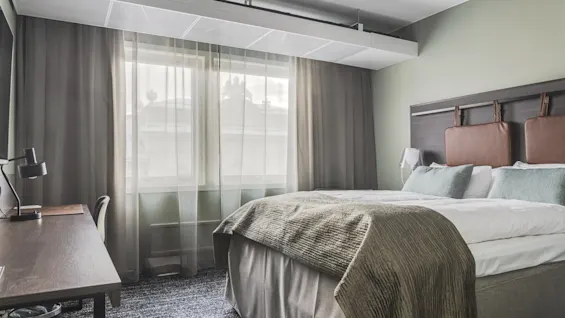Viihtyisä hotellihuone, jossa on mukava sänky, tyylikäs sängynpääty, toimiva työpöytä ja valoisa ikkuna verhoineen.
