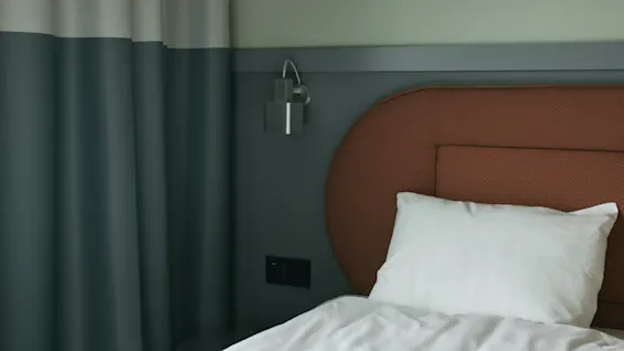 Ett mysigt och modernt hotellrum på Stopover Hotel Falkenberg, med en säng med vita lakan, en brun sänggavel och turkosa gardiner.