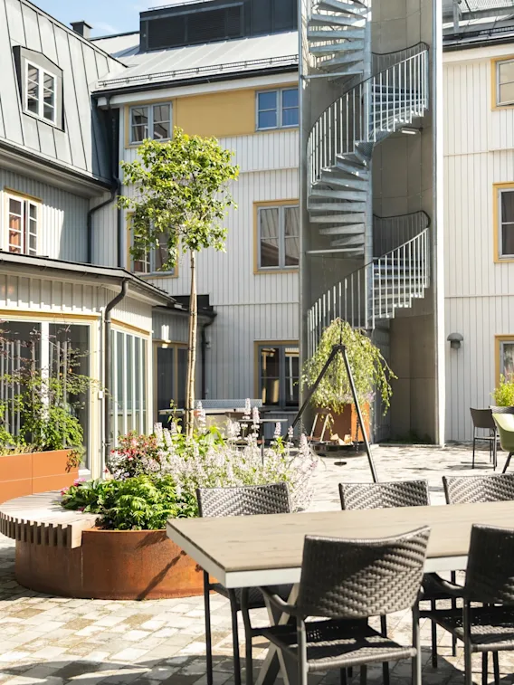 A sunny outdoor courtyard with various seating areas, lush plants, and a spiral staircase leading to upper floors.