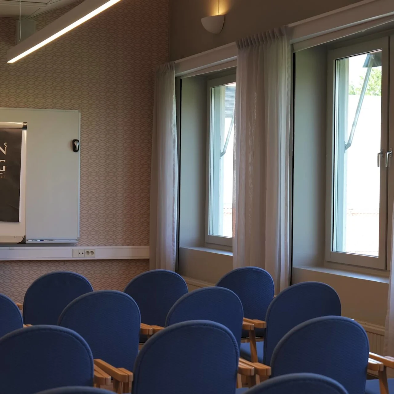 En flipchart viser "ARON BORG Konferenshotellet" og "Inblick, innebörd och ställtid" i et konferencerum med blå stole og vinduer med gennemsigtige gardiner.