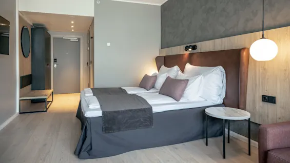 A bed is neatly made with white linens and gray blankets in a modern hotel room, featuring wood-paneled walls, a round pendant lamp, and a side table.