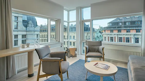 wooden panel wall background chairs sit near a coffee table with a magazine, framed by large windows offering views of adjacent historic buildings under a blue sky.