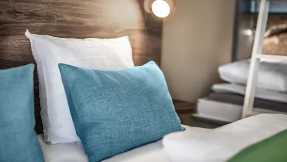 A bed with blue and white pillows sits neatly arranged. A soft light illuminates from a wall-mounted lamp in a cozy, modern room.
