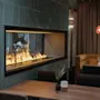 En modern eldstad med flammor brinnande stadigt är inbyggd i en kaklad vägg. Omkring den finns bord, stolar och en mysig bås i en varmt belyst, stilren restaurang.