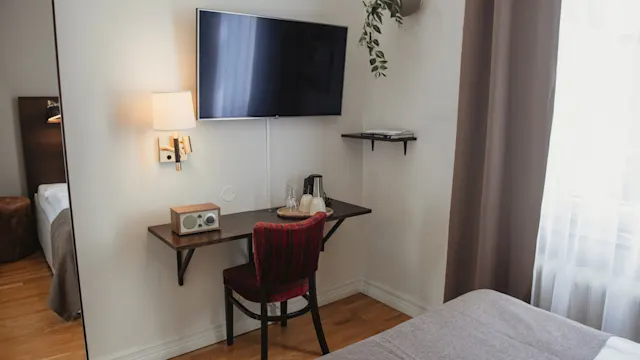 Ett litet skrivbord med en röd stol har en radio och vattenkokare under en väggmonterad TV. Ett närliggande fönster med flödande gardiner ljusar upp rummet med trägolv och ljusa väggar.
