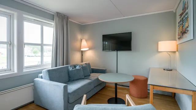 En grå sofa står i en stue med et rundt bord, lenestol og TV på veggen. To lamper lyser opp rommet, og et vindu slipper inn naturlig lys.