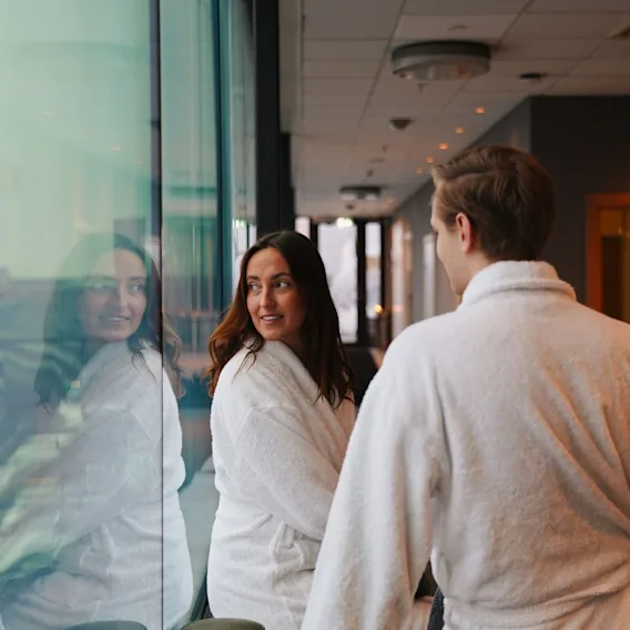 A man and woman wearing white bathrobes standing near a large window in a cozy spa relax area.
