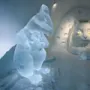 Isskulptur af en rotte, der holder ost, står i en snegrotte, overfor et stort katteansigt med lysende øjne, placeret i en struktureret snevæg.
