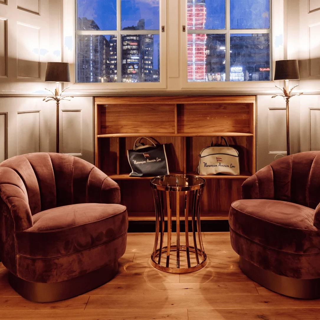 Two pink velvet chairs facing a glass-top table in a cozy room with wooden flooring. Shelves display two bags labeled "Norwegian America Line." A large window shows a cityscape at night.
