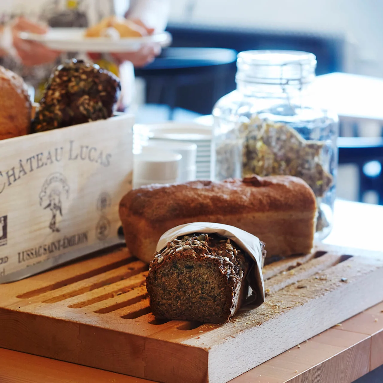 Bread on a wooden board, some in a basket, focus on a half-wrapped seeded loaf with blurred background.