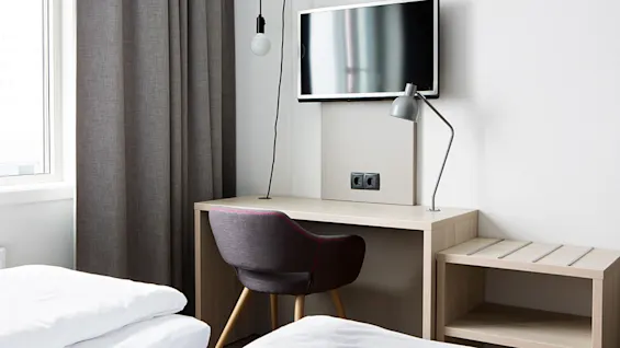 A minimalist hotel room with a desk, chair, bed, and mounted TV.