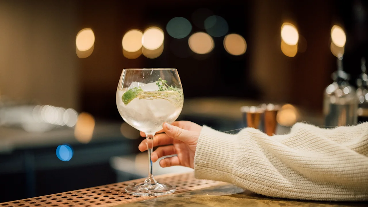 A hand holds a cocktail glass filled with a clear liquid, ice, and mint, set against a dimly lit bar with blurred warm lights in the background.