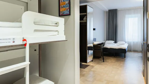 Våningssängar med säkerhetsetikett i ett modernt hotellrum med en snyggt bäddad enkelsäng, trägolv och ett skrivbord vid ett fönster med tunna gardiner. Text: "Max belastning 150 kg, Maximum load 150 kg."