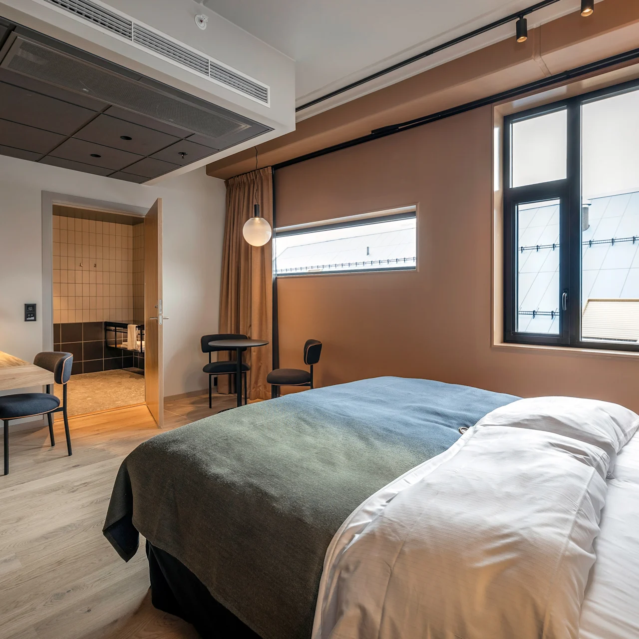 En pænt redt seng med grønt og hvidt sengetøj står i et moderne hotelværelse. Et skrivebord i træ, stole og en lampe fuldender rummets minimalistiske indretning ved siden af en åben badeværelsesdør.