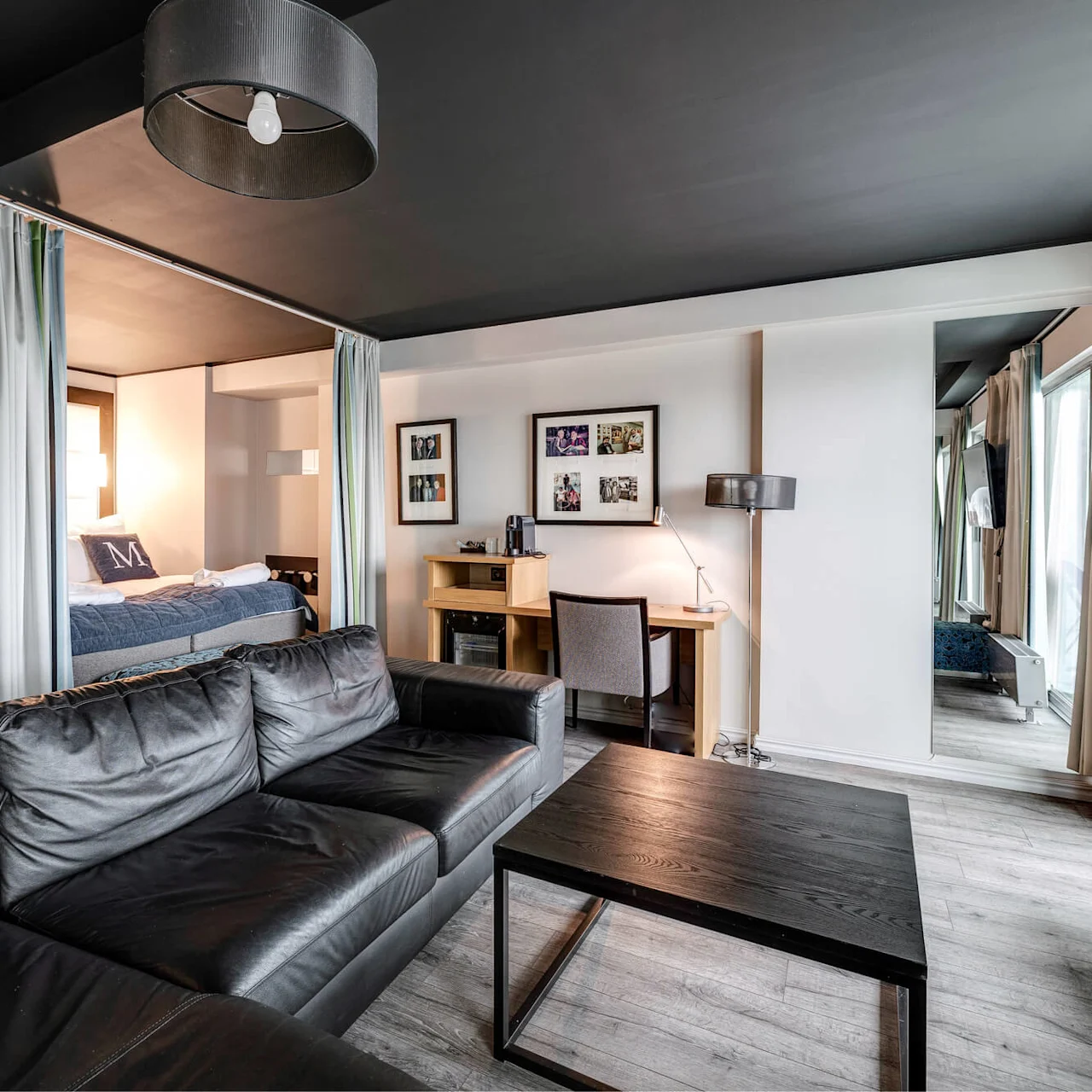 En svart lädersoffa vetter mot ett soffbord i ett modernt vardagsrum med grå väggar. En säng syns delvis bakom gardiner; ett skrivbord och foton pryder väggen.