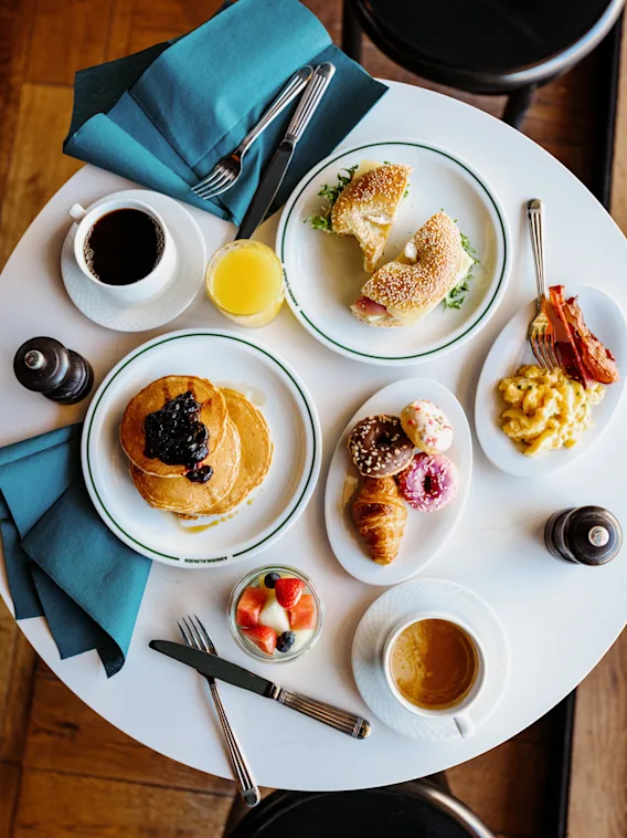 A vibrant breakfast spread on a white table, featuring pancakes, bagels, pastries, fruit, coffee, and juice. AMERIKALINJEN.