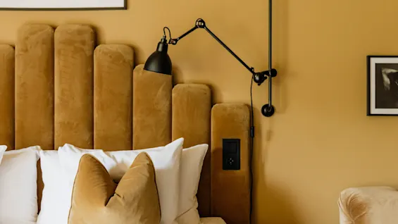 Et varmt og innbydende soverom med en plysj sennepsgul sengegavl, myke puter og en moderne svart leselampe.