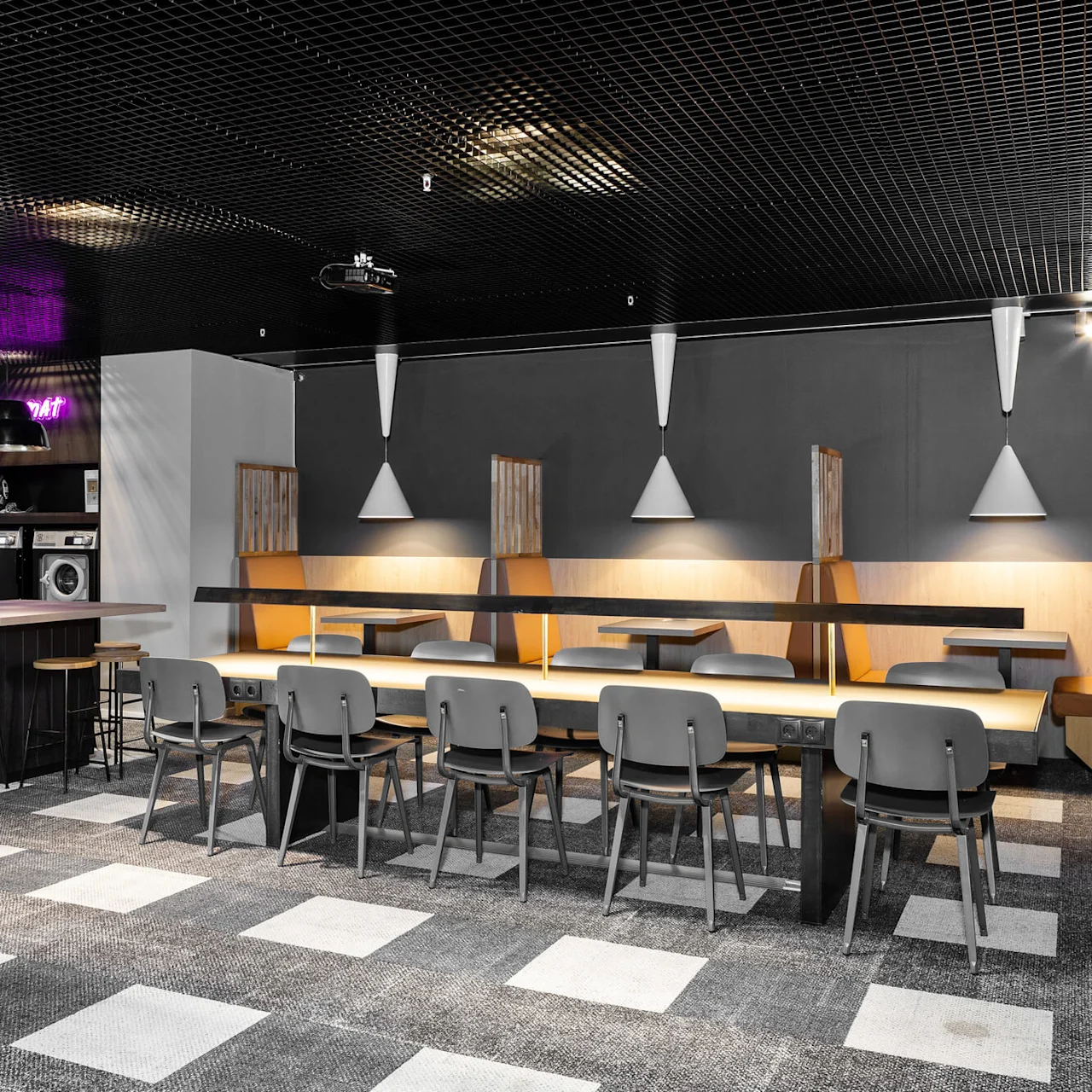A communal dining area with tables and chairs; washing machines and a neon sign reading "LAUNDRYMAT" are in the background. Modern lighting and decor create a relaxed atmosphere.