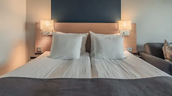 A double bed with white sheets and pillows is neatly arranged in a modern room. Two wall lamps illuminate the wooden headboard. A dark sofa is partially visible on the right.