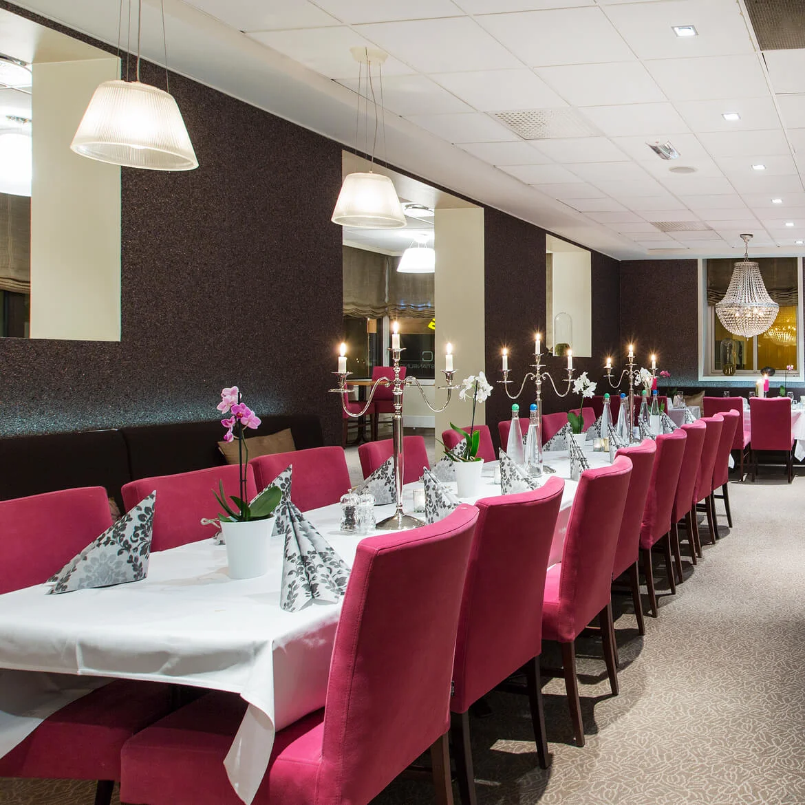 Spisebord er dekket med hvite duker, rosa stoler og lys, omgitt av et varmt opplyst, elegant restaurantinteriør med moderne dekor.