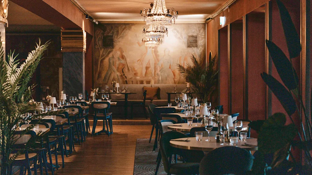 Bord dekket til middag; tomme, pyntet med glass og bestikk. Handling: Venter på gjester. Kontekst: Elegant restaurant med lysekroner, veggkunst og store planter, skaper en sofistikert atmosfære.