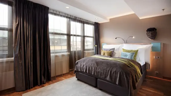 A cozy bed with pillows and a brown blanket is in a well-lit room with large windows and dark curtains; a light fixture and wall decoration add to the warm ambiance.