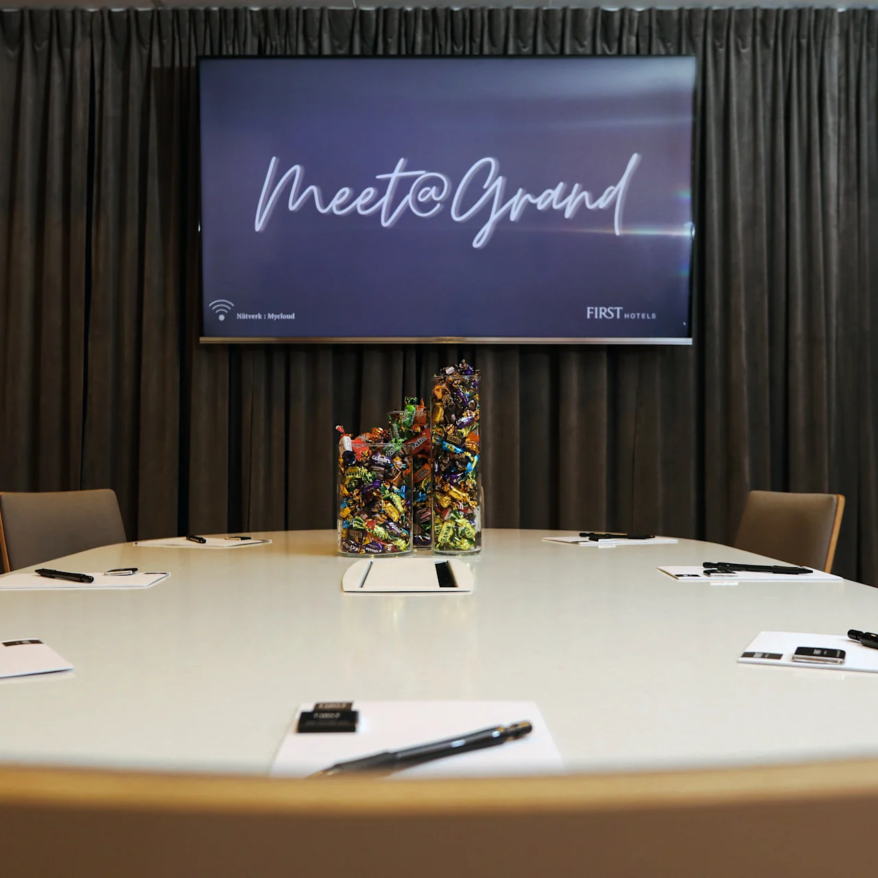 A conference room features a round table with notebooks and pens, centered with a colorful candy holder. A screen displays "Meet@Grand" against a curtained backdrop.