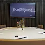 A conference room features a round table with notebooks and pens, centered with a colorful candy holder. A screen displays "Meet@Grand" against a curtained backdrop.