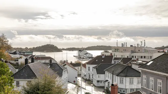 En charmig kuststad med vita och grå hus på en slingrande gata, med utsikt över lugnt vatten under en molnig himmel.