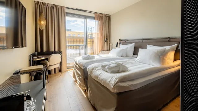 A neatly made twin bed sits in a tidy hotel room bathed in sunlight, with an open balcony door and urban view outside.