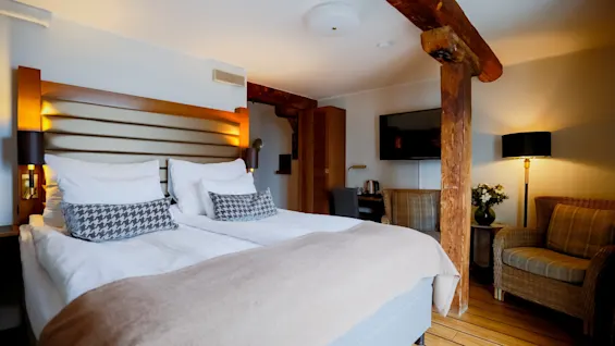 Et hyggeligt hotelværelse med en stor seng med houndstooth-puder, et vægmonteret TV og rustikke træbjælker.