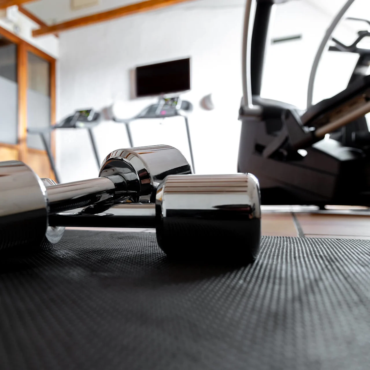 A chrome dumbbell rests on a black mat; treadmills and exercise equipment in the background of a well-lit gym.