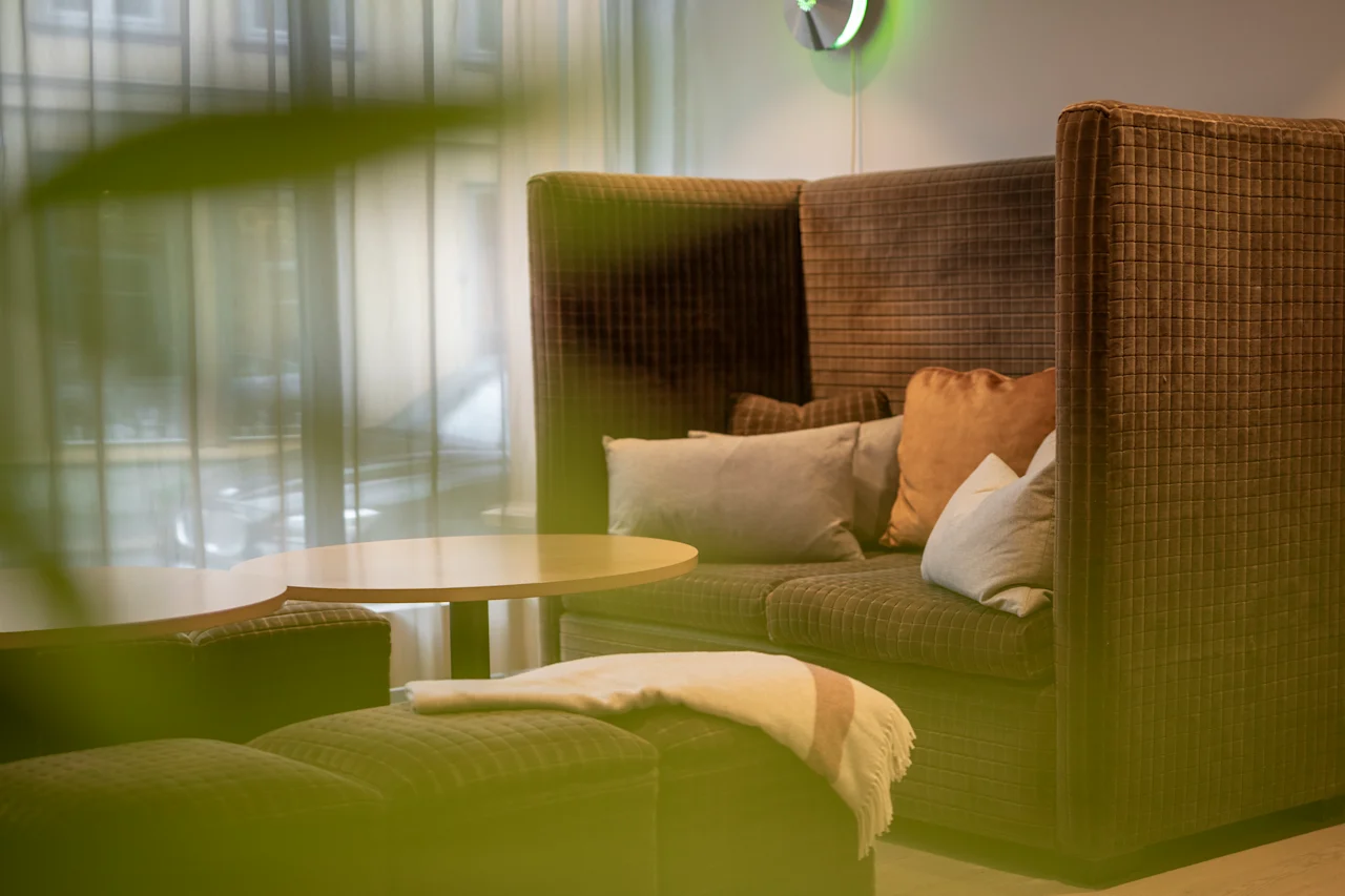 En brun, polstret sofa med puter står ved siden av et rundt bord i et mykt opplyst rom med tyllgardiner og en grønn plante i forgrunnen.