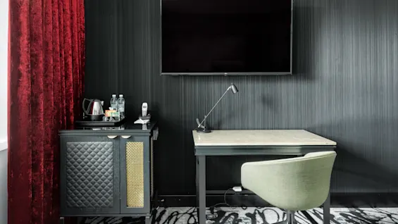 A modern desk with a table lamp and chair against a textured wall, flanked by thick red curtains and a flat-screen TV above.