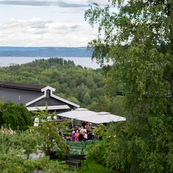 Oversiktsbilde av gjester på en utendørsterrasse under store hvite parasoller omgitt av grønn natur og en fjord.