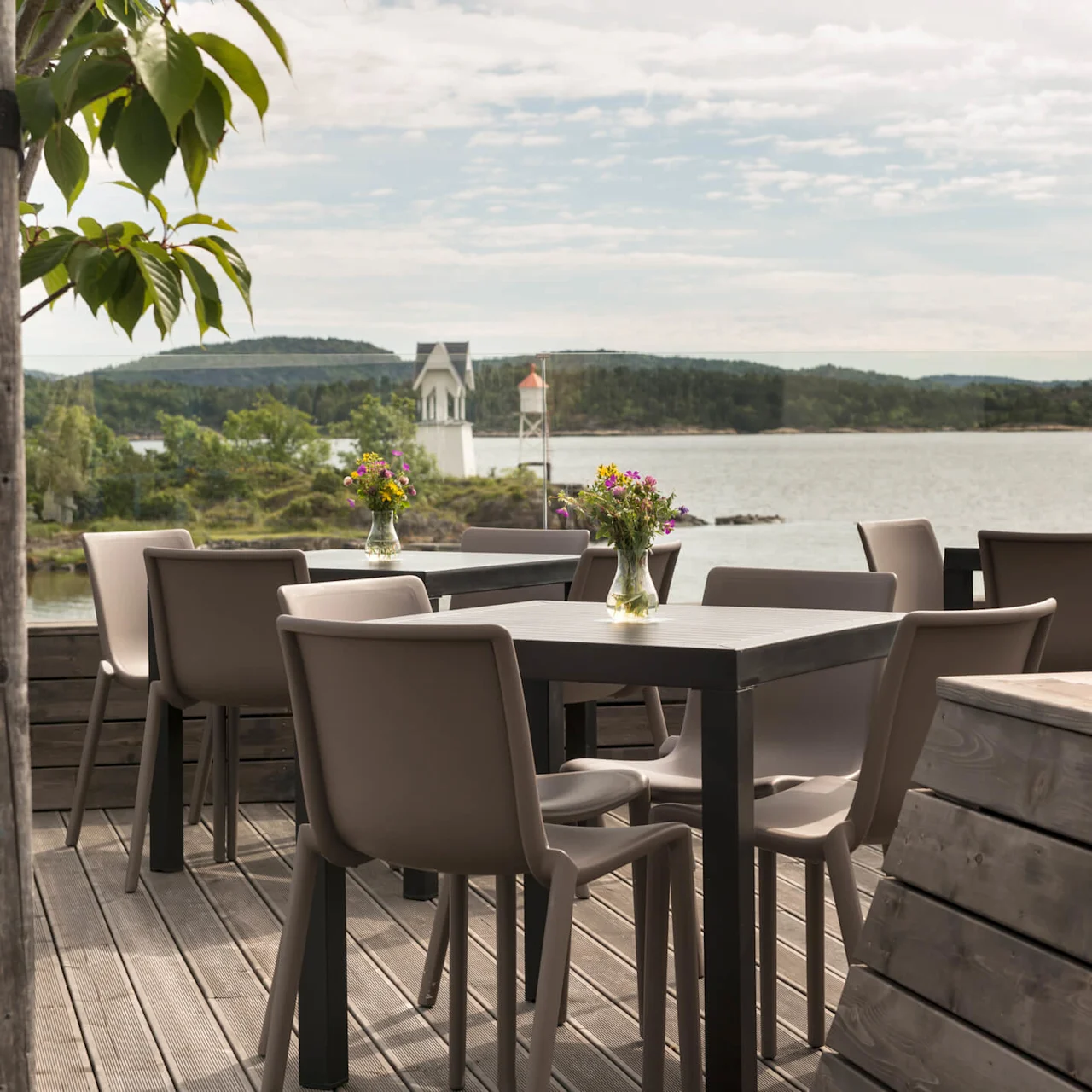 Tables arranged on a wooden deck hold vases of colorful flowers; green trees and a calm lake with distant hills form the serene backdrop.