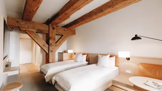Ljust hotellrum med enkelsängar, ljusa trädetaljer och rustika synliga takbjälkar i trä.
