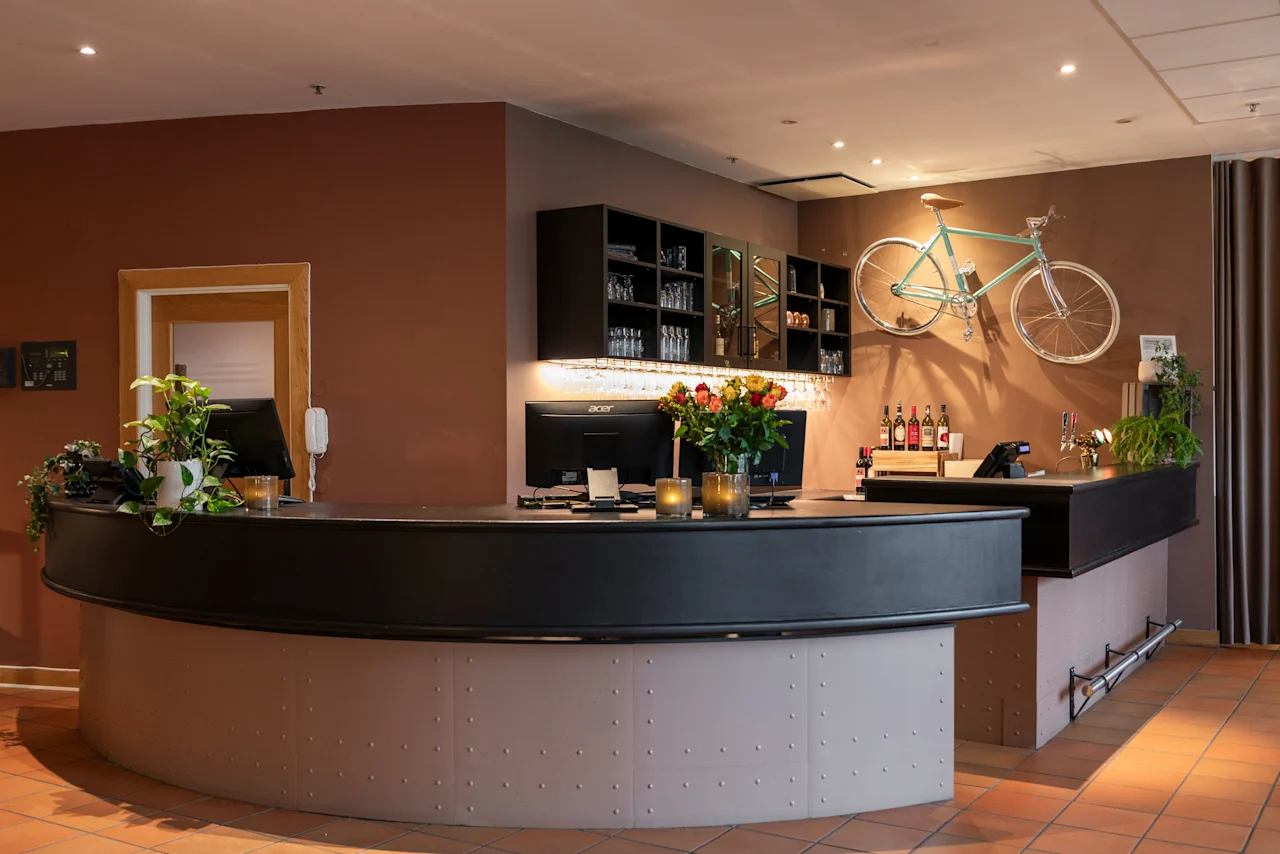 A stylish hotel reception and bar area with a curved counter, monitors, flowers, and a bicycle mounted on a brown wall.
