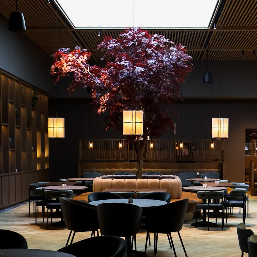 A tree with red leaves stands indoors, surrounded by modern round tables and chairs. Warm pendant lights hang from a wooden ceiling in a stylish dining area.