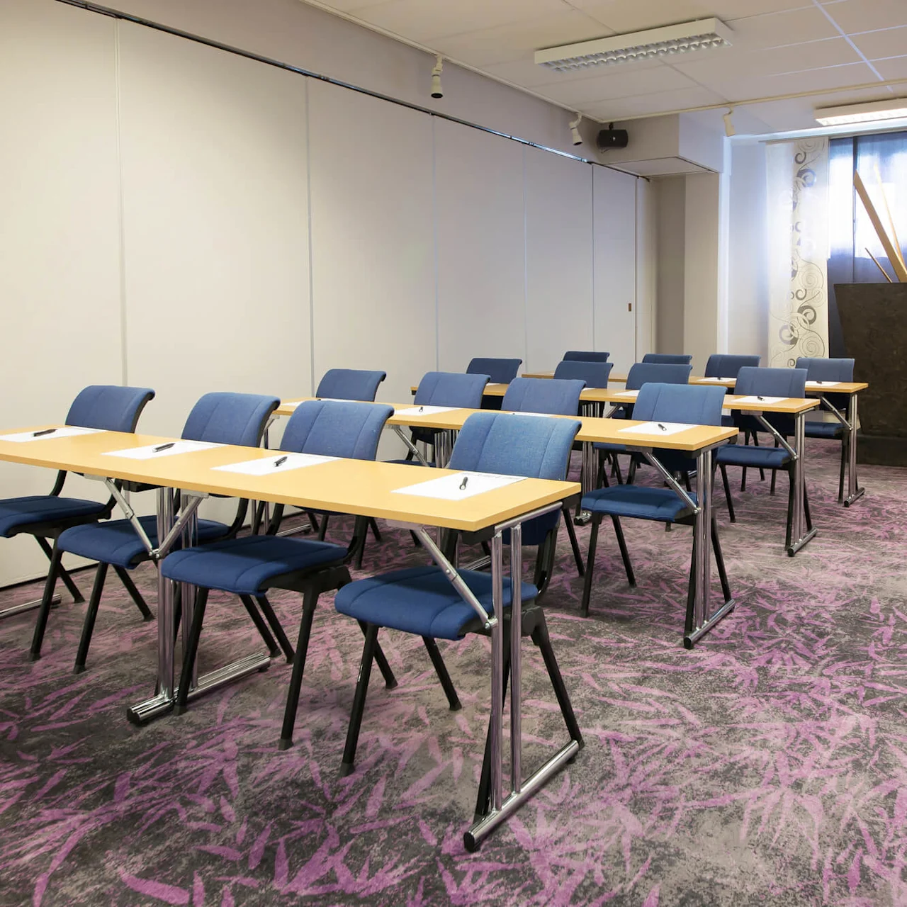 Rader av bord med papper och pennor, förberedda för deltagare i ett seminarierum med dekorativa gardiner och konst.