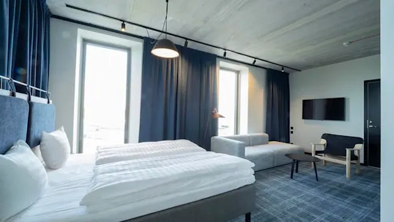 A modern hotel room with two beds, a seating area, and large windows, offering a comfortable stay.