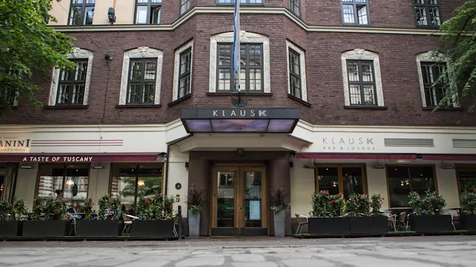 Bygningsfasade med "KLAUS K"-skilt over inngangen. Gateomgivelse inkludert en kafé med rød markise merket "A TASTE OF TUSCANY" og uteservering omgitt av planter.