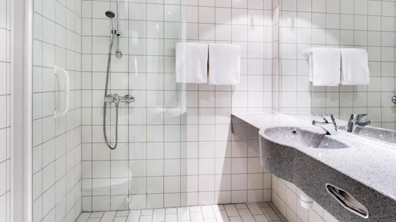 Bruser med glasdør i flisebelagt badeværelse, med håndbruser og tilstødende vask med to haner. To hvide håndklæder hænger på nærliggende holder.