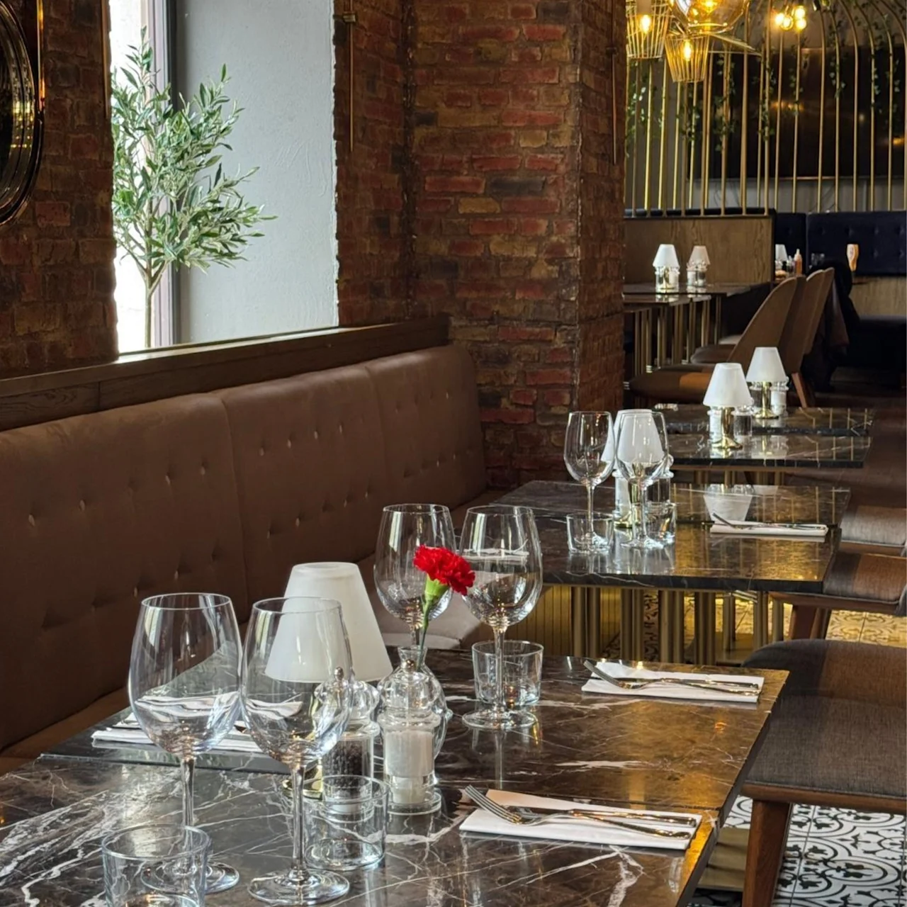 Et restaurantbord dækket med vinglas, servietter og en rød blomst i en vase. Placeret i et varmt oplyst, elegant spisestue med murstensvægge og hængende lys.