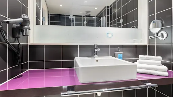 A modern bathroom sink with a large mirror, black tiled walls, and white towels. There's a hairdryer on the left and a text sticker saying "DRINK MORE WATER".