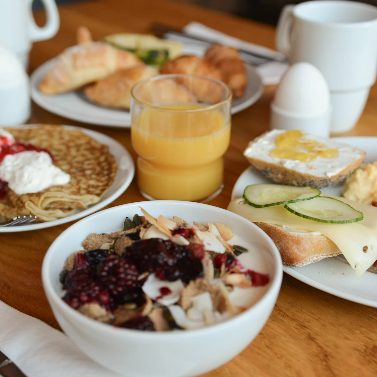 Skåle og tallerkener med forskellige morgenmadsprodukter, herunder pandekager med fløde, korn med bær, røræg, skinke, skåret agurk på brød, croissanter og et glas appelsinjuice på et træbord.