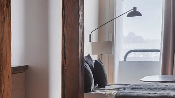 A cozy bed with dark cushions and blanket is situated near a large window, illuminated by a modern floor lamp, framed by wooden beams. 