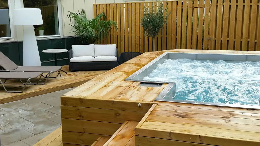Boblebad, bobler energisk, plassert på en treterrasse. Omgitt av solsenger, sofa, bord og potteplanter mot et tregjerde.