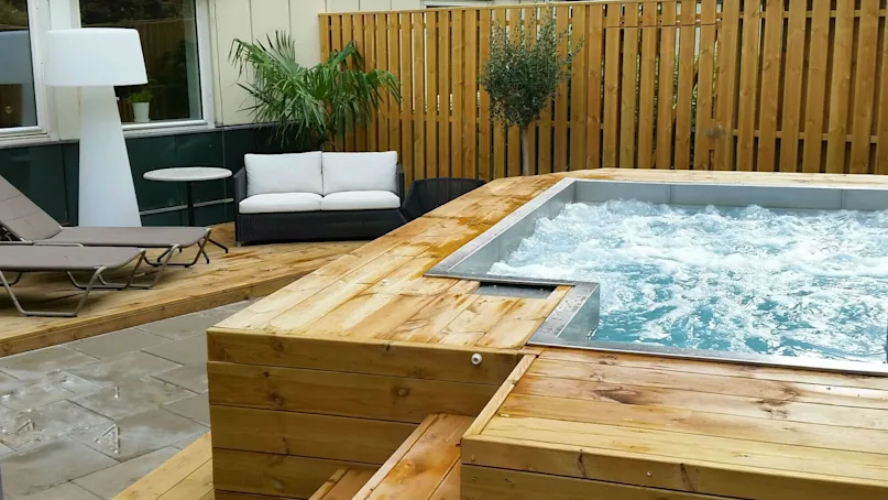 Jacuzzi, bubbling energetically, situated on a wooden deck. Surrounded by lounge chairs, a sofa, a table, and potted plants against a wooden fence.
